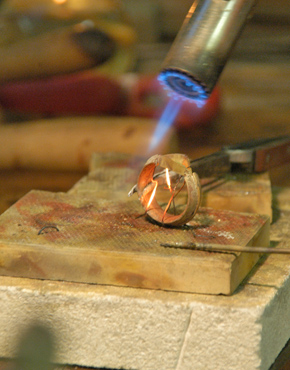 Bild auf dem ein Ring zu sehen ist, der mit einer Pinzette über einem Lötblock gehalten und von einem Lötkolben, für die Reparatur, erhitzt wird.